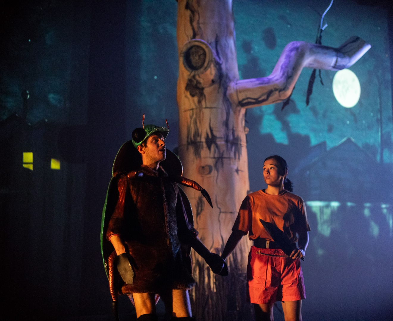 A man dressed as a beetle holding hands with a woman against a backdrop of a moonlit night with a gum tree