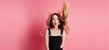 Photo of comedian Madeleine Stewart against a pink background. She is wearing black overalls and her long blonde hair is being comically blown upwards.