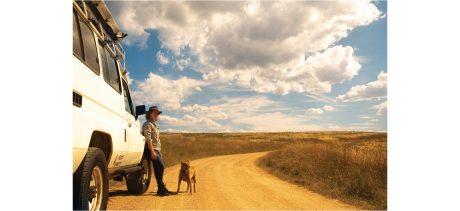 A white land rover is parked in the foreground of the image on a yellow dirt road that stretches to a light blue sky horizon with white clouds. A woman with an akubra hat rests against the car, there is an rusty coloured kelpie dog at her feet. 
