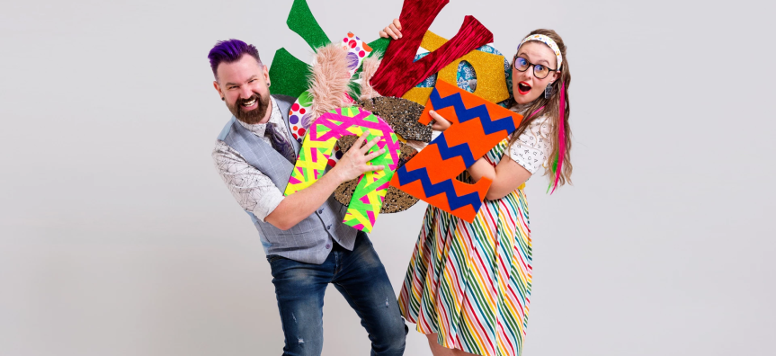 A man in a grey vest and jeans with a woman in a colorful skirt and white t-shirt hold onto an assortment of colourful cardboard letters - they look excited. 