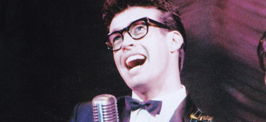 Close up image of performer Scot Robin as Buddy Holly, singing into a vintage microphone. He wears a suit and bowtie and thick rimmed black glasses. He looks happy.