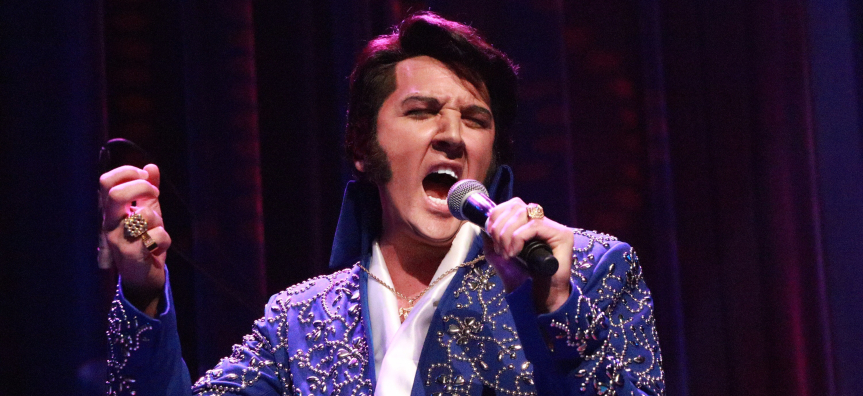 Performer Mark Anthony pictured, dressed as Elvis with a white open-necked shirt and a purple jacket threaded with gold patterns. He is holding the mic stand in one hand and singing passionately into the mic in the other. He rocks a classic Elvis hair do flipped over his forehead.