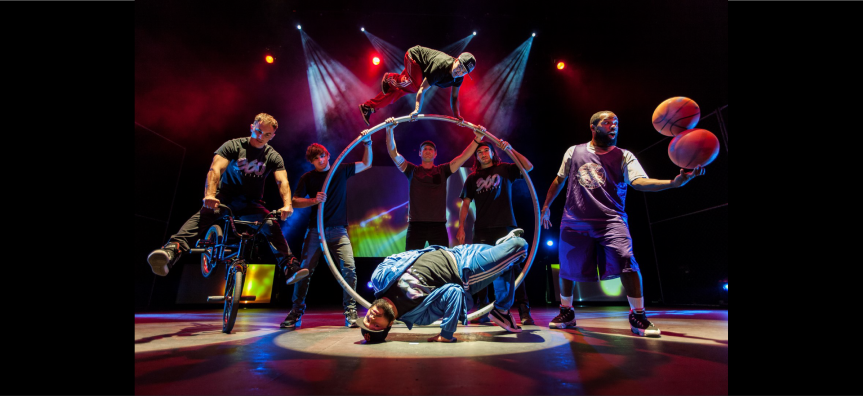 360 Allstars crew performing acrobatics. One man juggles two basketballs in one hand, another spins on a BMX, several crew hold a rotating silver aerial hoop while another breakdances in the foreground.