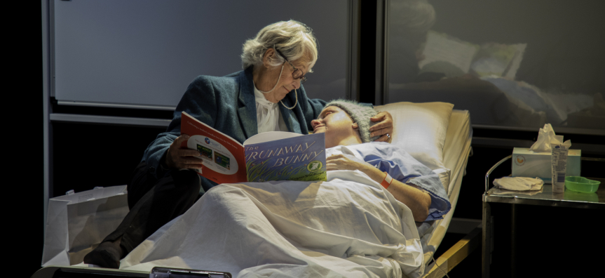 Image of a woman lying in a hospital bed talking to an older woman. The older woman is reading to her from a children's book.