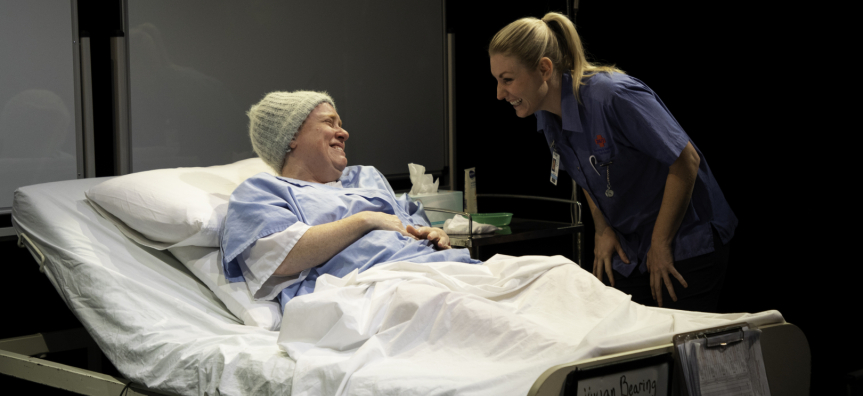 Image of a woman lying in a hospital bed talking to another woman, her nurse. 