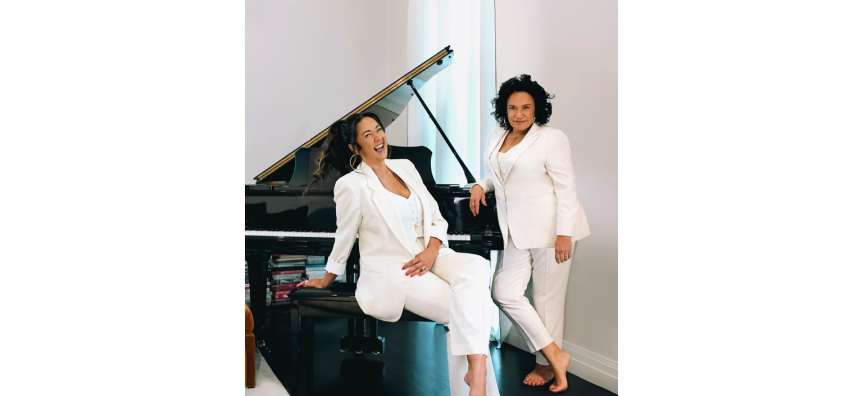 Singers Vika & Linda Bull sit in front of a grand piano. They are wearing white suits and smiling happily