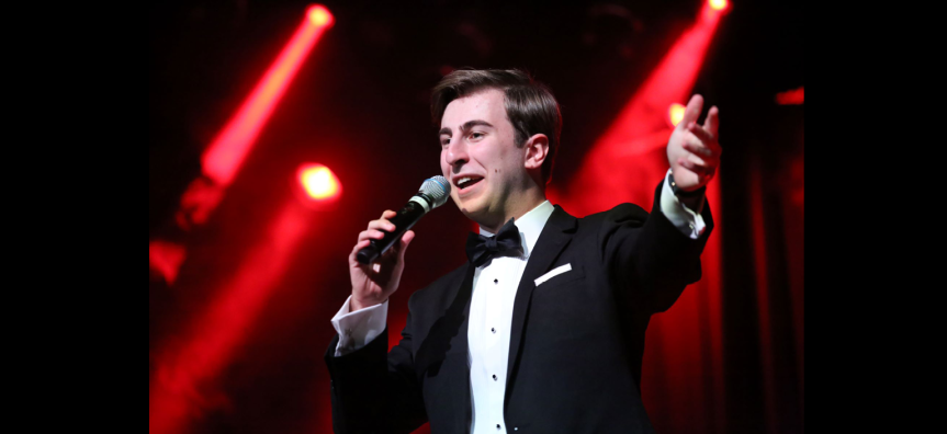 A young man sings into a microphone, he wears a tuxedo. 