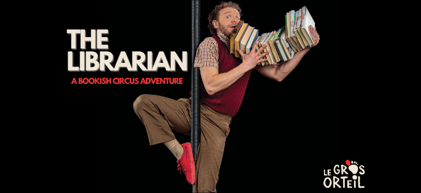 A man with curly brown hair holds onto a precarious stack of books in his arms, while looping a foot around a tall pole
