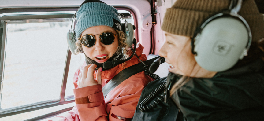 Two women smile at one another in a helicopter. They are flying over snowy mountains and are wearing beanies and headsets