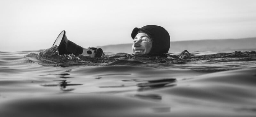 A woman in a thick wetsuit with a waterproof camera is seen smiling and bobbing in the waves. The photo is in black and white. 