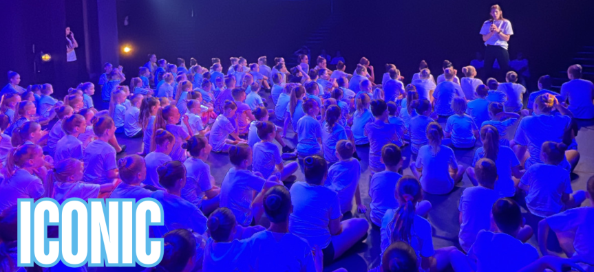 A large crowd of young dancers sit on the floor watching a stage with a woman speaking to them. The word Iconic is written in the left-hand corner in bold