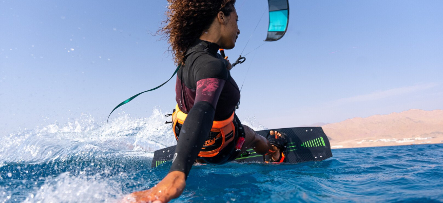 A woman seen from behind kitesurfing on the ocean, her hair is brown and curly and flies wildly in the wind