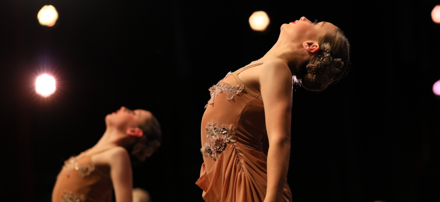 Two young female dancers are posed against a black backdrop with theatrical lights shining on them 