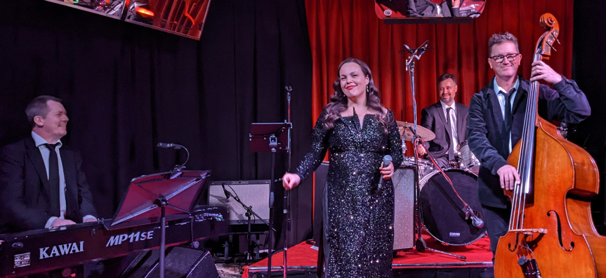 A female singer in black dress stands between two men in suits, one playing piano and one playing the cello. Performers are smiling as they sing and play their instruments.