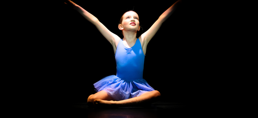 A young girl in a blue leotard sits on a stage with her hands raised to the sky smiling. 