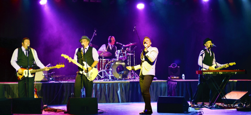 Four men in suits are positioned ina line on stage playing guitars and singing. Purple lights are shining on them from the background. 