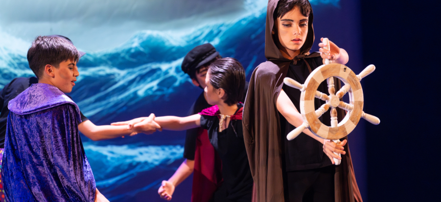 A young boy in brown robes pretends to steer a ship using a ship wheel while 4 boys in the background clasp hands 