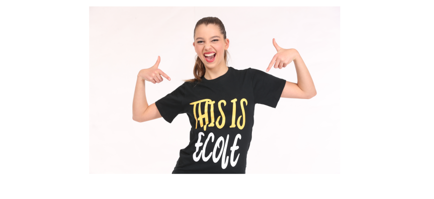 A woman standing against a white background smiles and points to her shirt that says 'this is ecole'