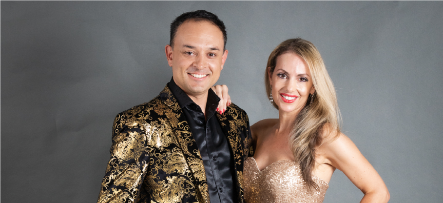 Two performers smiling at the camera, a man in a glitzy jacket and a woman with long hair in a sparkly evening gown