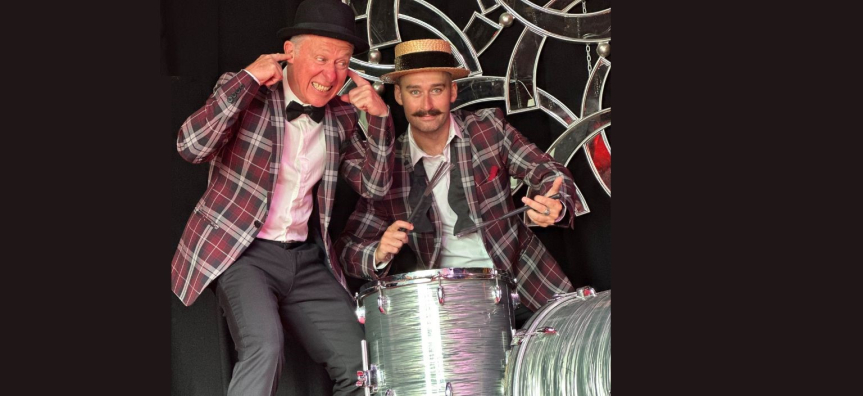 I man in a plaid blazer and straw hat playing the drums as another man also in a plaid blazer grimacing and puts his fingers in his ears.