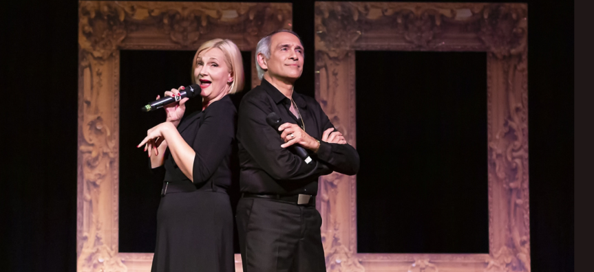 A woman in a black dress and a man in a black suit stand back to back on stage, she is singing into a microphone