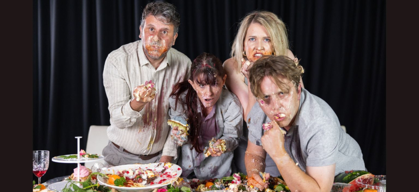 Four people leaning over a table full of food, the food is all over them, their clothes and faces.