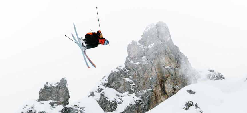 Aerial skiier mid jump, skis are crossed and there are snow covered mountains in the background