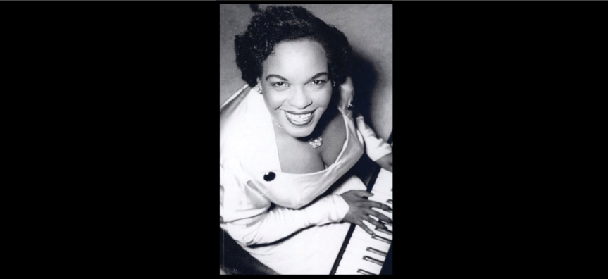 A photo of legendary musician Winnie Atwell at her piano smiling broadly at the camera. The photo is in black and white.