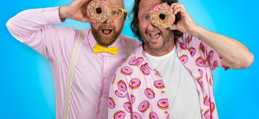 A smiling man in a pink shirt with yellow and white striped braces holds a pink doughnut with sprinkles up to his eye and another smiling man in a pink shirt covered in pink doughnuts holds a pink doughnut with sprinkles up to his eye.