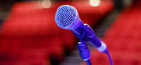 A closeup of a microphone, in the background are empty red theatre chairs