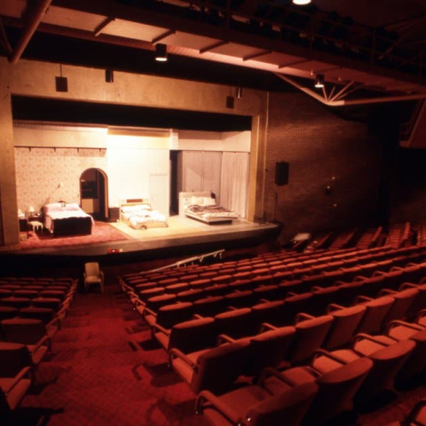 Image of the interior of a theatre with rows of seating and a lit stage with a bedroom scene in place